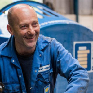 A man smiling in a workshop in his blue work overalls while people work behind him